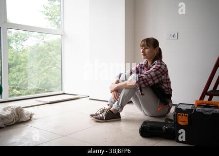 Female repair worker in casual shirt sitting on floor beside ladder and toolbox during home renovation. Concept of DIY repair, maintenance, and improv Stock Photo