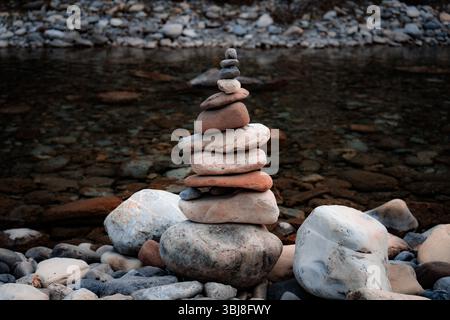 A balanced stack of smooth river rocks, arranged in ascending size, stands on a rocky shoreline Stock Photo
