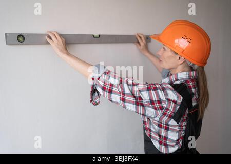 Female construction worker wearing orange hard hat measures the walls a spirit level. Stock Photo