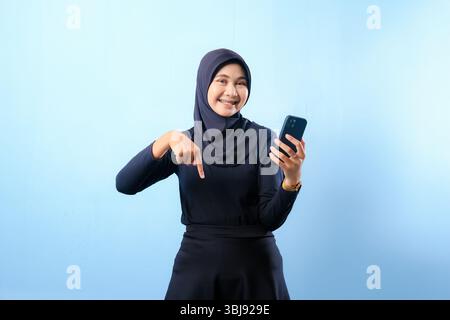 A woman wearing a black hijab is holding a cell phone and pointing to the camera. She is smiling and she is happy Stock Photo