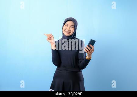 A woman wearing a black hijab is holding a cell phone and pointing to the camera. She is smiling and she is happy Stock Photo
