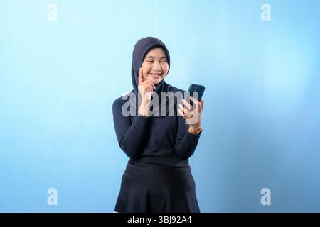 A woman wearing a black hijab is holding a cell phone and pointing to the camera. She is smiling and she is happy Stock Photo