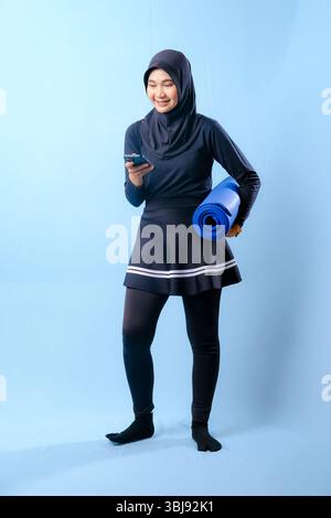 A woman in a black skirt and black pants is holding a blue yoga mat. She is looking at her cell phone Stock Photo
