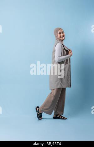 A woman wearing a grey vest and a white shirt is smiling at the camera. She is wearing a headscarf and has her arms crossed Stock Photo