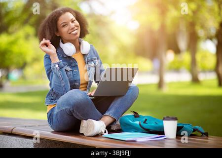 Black student woman sitting outdoors on campus bench with laptop on lap Stock Photo