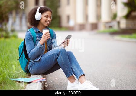 Happy Black Student Woman Using Smartphone And Drinking Coffee Outdoors Stock Photo
