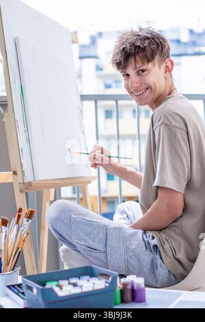 Cheerful, cute teenager at an easel learning to draw with colored paints and brushes on an easel, smiling sincerely while looking into the camera. Hig Stock Photo