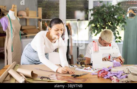 Attentive young man tailor sewing textile using sewing machine and female assistant drawing pattern on sheet of paper using curves in fashionable Stock Photo