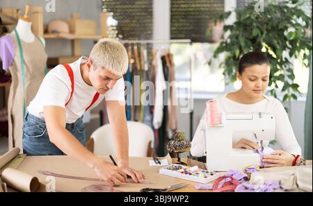 Attentive woman tailor sewing textile using sewing machine and young male assistant drawing pattern on sheet of paper using curves in fashionable Stock Photo