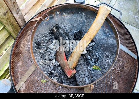 Close-up view of a rusty fire pit with a charred log and an axe embedded in it, surrounded by ash and burnt wood remains. Stock Photo