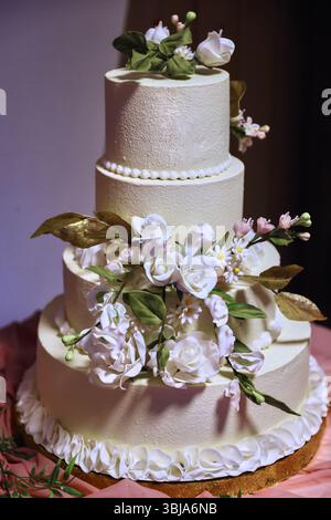 Elegant four-tiered white wedding cake decorated with beautiful white flowers and green leaves. Luxury dessert for a celebration or marriage event Stock Photo