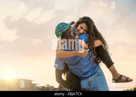 A happy diverse couple in silhouette against a golden sunset on a beach. The man gives the woman a piggyback ride in a romantic and carefree moment du Stock Photo