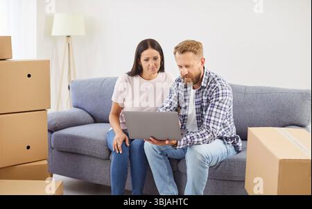 Young couple sitting on sofa in new apartments using laptop together with unpacking cardboard boxes Stock Photo