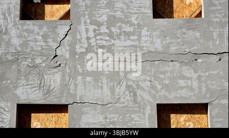 Close-up of gray concrete wall with deep horizontal and vertical cracks, rough surface texture, and rectangular window openings, revealing plywood Stock Photo