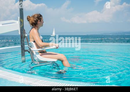 Young woman with a disability sitting in a pool chair lift above the poolside overlooking the marine horizon and enjoying a sunny day by the sea. Stock Photo