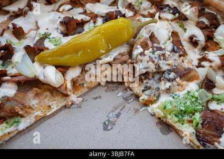A half-devoured doner kebab pizza in close-up, creamy sauce dripping, with a tangy pickled pepper, still tempting in its box. Stock Photo