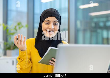 A young woman in a hijab smiles broadly while looking at a laptop, holding a notebook and pen in an office setting. Stock Photo