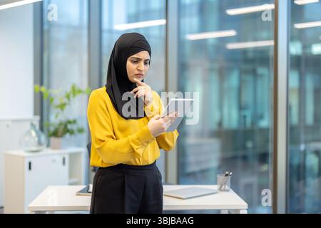 A thoughtful businesswoman in a hijab reviews information on a tablet inside a modern office setting, contemplating her next action. Stock Photo