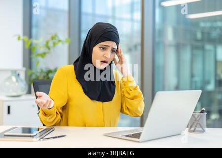 A woman wearing a hijab looks stressed while working on her laptop in an office setting. Stock Photo
