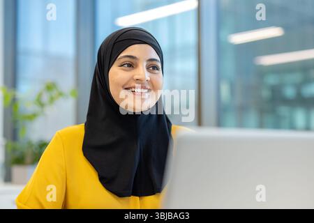 A smiling woman wearing a black hijab looks towards the right in a modern office setting. She is wearing a yellow shirt. Stock Photo