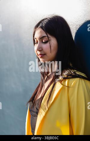 Young woman wearing a yellow blazer stands quietly with eyes closed, leaning against a sunlit blue wall, creating a calm and introspective outdoor por Stock Photo