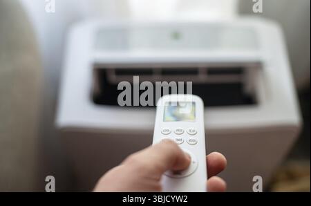 Hand holding remote control aimed at floor standing mobile air conditioner Stock Photo