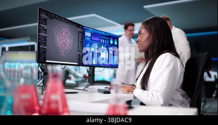 Young Scientist Conducts Research In Manufacturing Lab, Analyzing Data On Screens. A Diverse Group Works Together Stock Photo