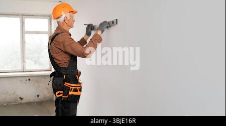 Elderly handyman. Senior builder in uniform measuring the wall using spirit level and making marks on wall Stock Photo