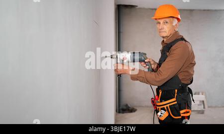 House renovation concept. Elderly construction worker in yellow hardhat. Builder or handyman works on renovation house using electric drill drills Stock Photo
