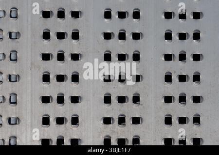 Close-up of a brushed metal surface with a repeating pattern of capsule-shaped holes. Light reflecting on the metallic material. Stock Photo