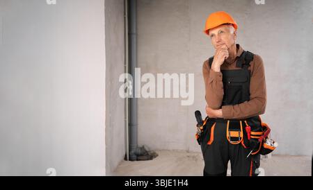 Senior builder on construction site standing wearing safety helmet thinking an idea while looking away. Elderly building worker Stock Photo