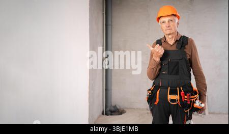 Elderly mature male builder in safety helmet say look there. Pointing thumb aside at blank space. profession, construction and building concept Stock Photo