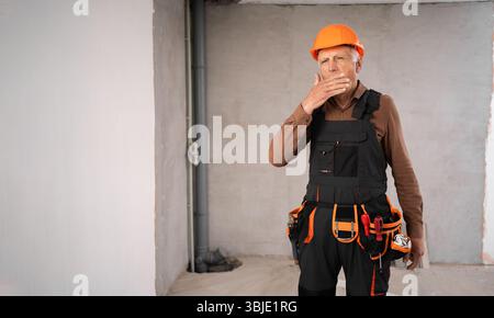 tired yawning sleepy pensioner builder construction site worker in protective suit and overalls at work place Stock Photo