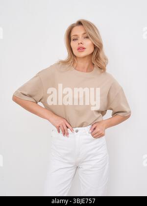 Blonde woman with wavy hair, wearing a beige short-sleeve top and white pants, poses confidently with hands on hips, gazing forward. Casual fashion Stock Photo