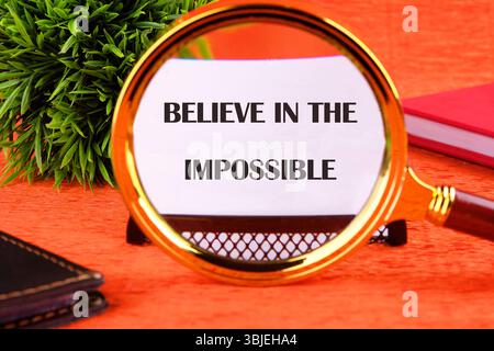 Motivational quote. BELIEVE IN THE IMPOSSIBLE text focuses through a magnifying glass on a business card on an orange background. Stock Photo
