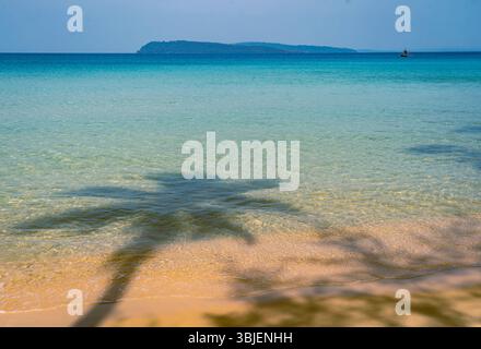 A serene beach scene featuring clear turquoise waters, soft sandy shore, and a palm tree casting a shadow on the sand. An island is visible in the dis Stock Photo