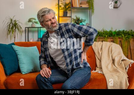 Tired senior bearded man suffering from back pain sitting on sofa in living room at home. Sad old male massages painful lower back muscles. Caucasian mature guy exhausted overworked in apartment. Stock Photo