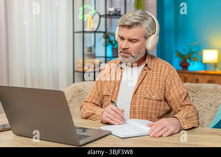 Caucasian mature man sitting at home table listening to podcast or online stream on laptop with headphones. Guy takes making notes into notebook, interested engaged in content, learning education Stock Photo