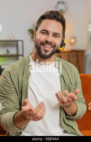 Confident young Caucasian man sitting on sofa at home wearing headset, freelance worker, call center, support service operator helpline, having talk with client or colleague, communication support. Stock Photo