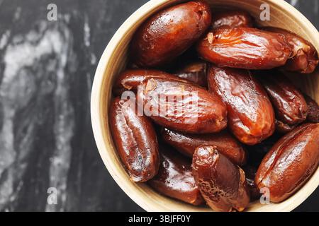 Dates in wooden bowl. Sweet dried fruit. Organic healthy snack. Natural sugar source. Brown glossy texture. Closeup on black marble. Stock Photo