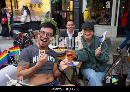 Three people, two men and one woman, are seated at an outdoor cafe table. They are holding small rainbow flags and stickers.  The scene is set on a ci Stock Photo