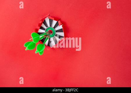 Three green darts in the middle of a target board, thru a ripped hole on red paper background, with copy space for text or product Stock Photo