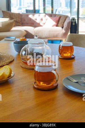 Glass teapot and cups with hot tea and lemon on wooden table in bright living room. Warm light and cozy interior. Concept of wellness and relaxation Stock Photo