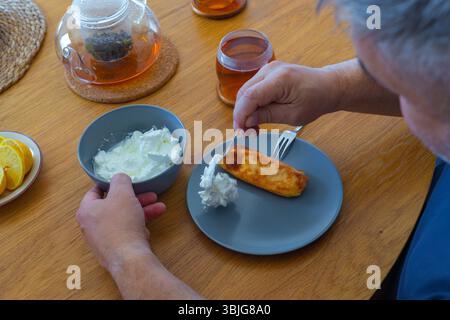 Close-up of man preparing to eat pancake with sour cream, glass of tea, and lemon slices on cozy wooden table at home. Casual morning atmosphere Stock Photo