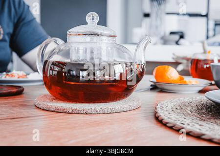 Glass teapot with brewed tea on a cozy kitchen table. Plate with fresh orange slices in the background. Warm indoor scene with natural light Stock Photo