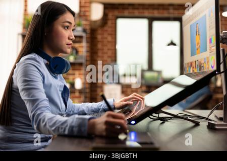 Asian graphic designer holding stylus pen, using digital art pad to modify images on computer screen. Skilled woman focused on retouching photos and refining design details for content production. Stock Photo