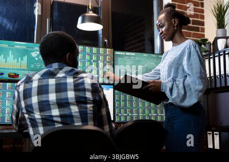 Two financial analysts collaborate at desk in office, reviewing stock market information on multiple monitors at night. Female manager with a clipboard, stands and explains forex trading to employee. Stock Photo