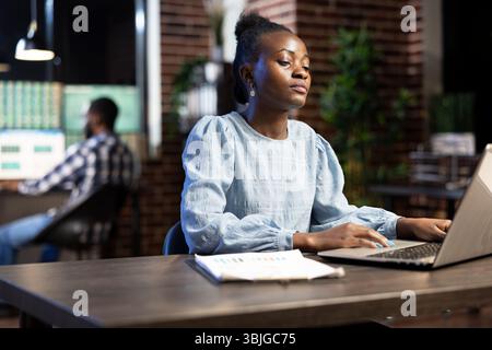Young businesswoman with clipboard nearby, working late at night, analyzing financial data on her laptop. A blurred male coworker in the background focuses on multiple monitors. Stock Photo