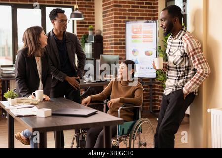 Multiethnic colleagues gather around a desk, taking a break and sharing stories in a relaxed office setting. Diverse group of employees stands, chatting with their asian coworker in a wheelchair. Stock Photo