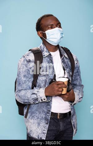 African american man in denim jacket and face mask holds his dslr camera and coffee cup, ready to meet his photography clients. Young photographer gazes upward while standing against isolated backdrop. Stock Photo